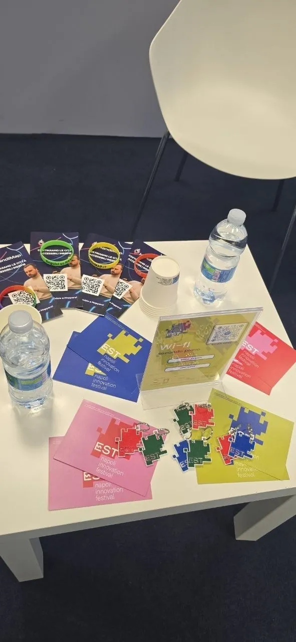 Tavolo dei relatori con braccialetti di HandiMap, cartoline di EST, pezzi di puzzle colorati e bottiglie d'acqua