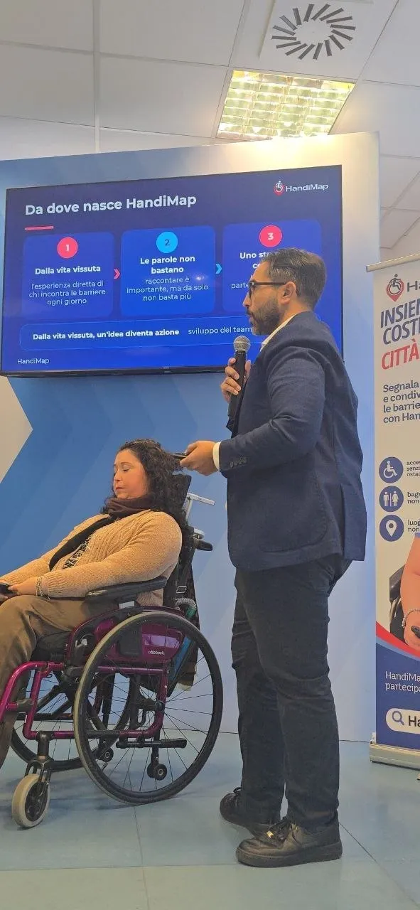 Manilla in carrozzina e Arcangelo con il microfono presentano HandiMap davanti allo schermo con la slide «Da dove nasce HandiMap»