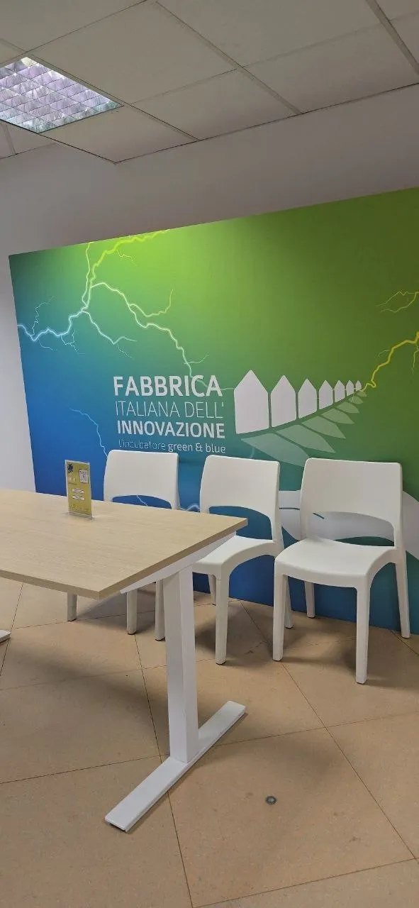 Parete verde con la scritta «Fabbrica Italiana dell'Innovazione», con tavolo e sedie bianche